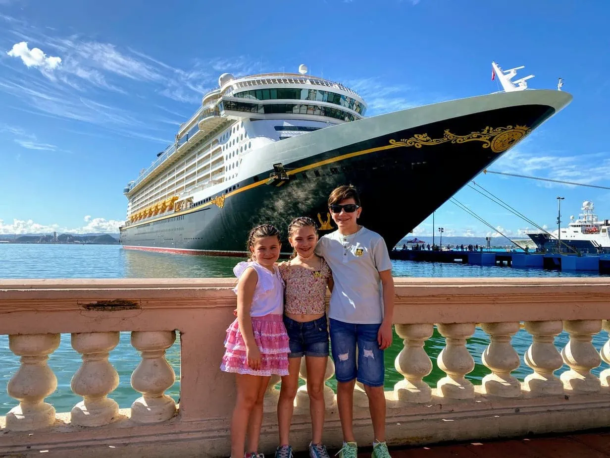 Kids standing in front of large Disney Cruise ship docked in blue seas