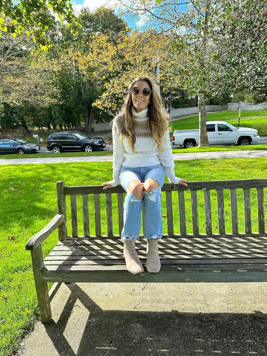 Brianne wearing the perfect fall outfit in a comfy sweater and denim jeans while sitting on NYC park bench