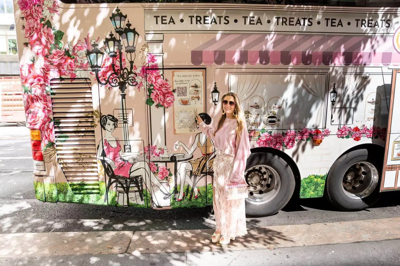 Brianne in all pink and floral ensemble standing in front of door of the Tea Around Town Bus 