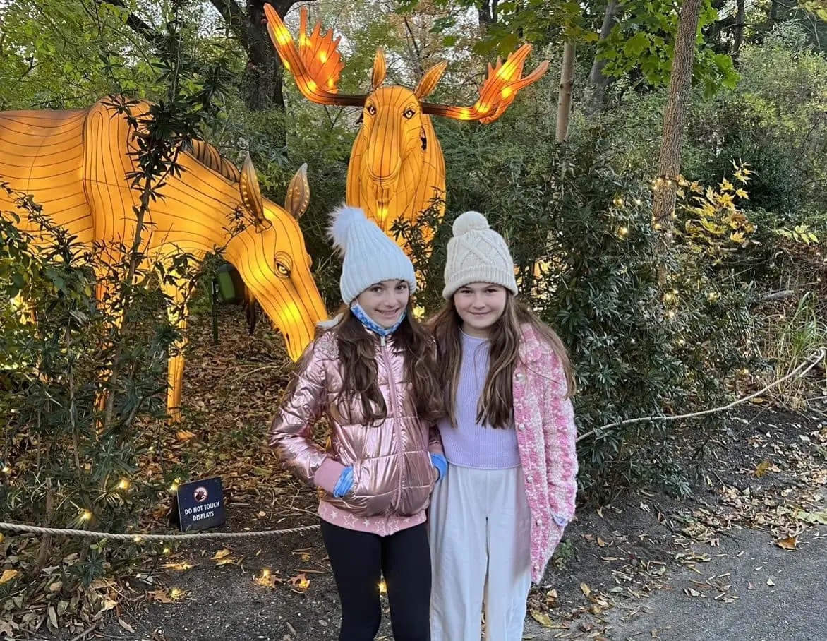 Daughters in cute white pom-pom winter hats smiling in front of NYC Bronx Zoo's holiday light display of reindeer in November 